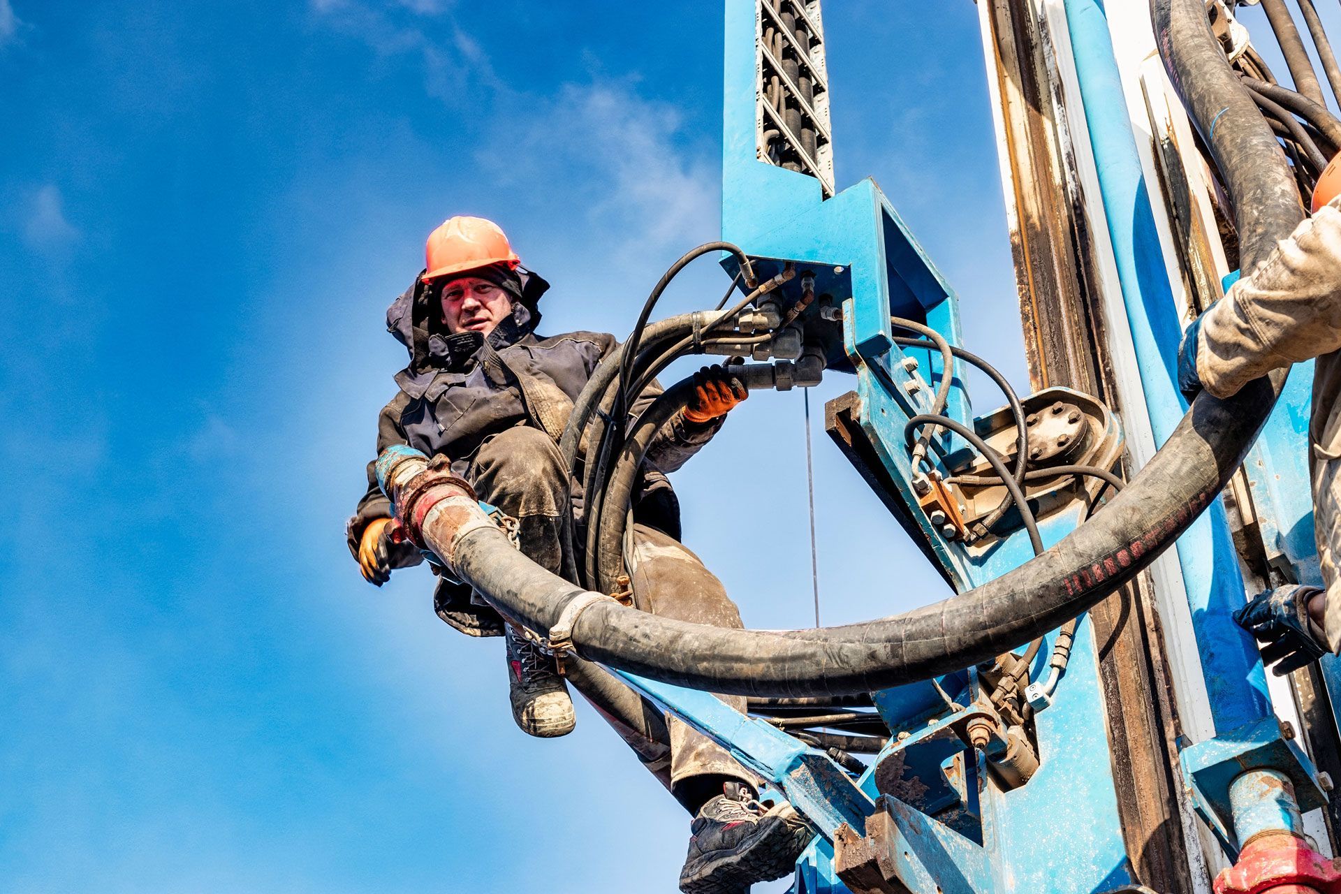 Worker in hard hat adjusts hoses on a blue drilling rig against a bright blue sky.