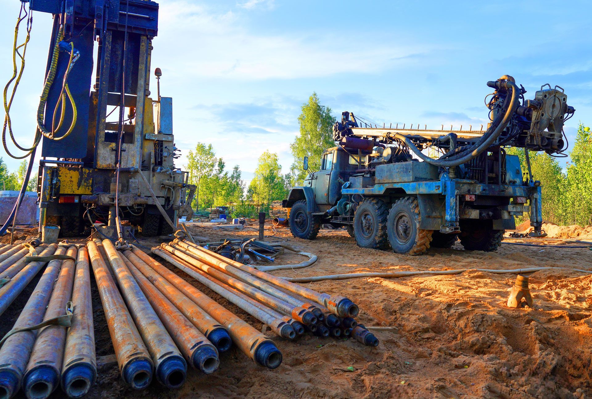 Drilling rig on a muddy worksite with pipes laid out; trees and sky in the background.