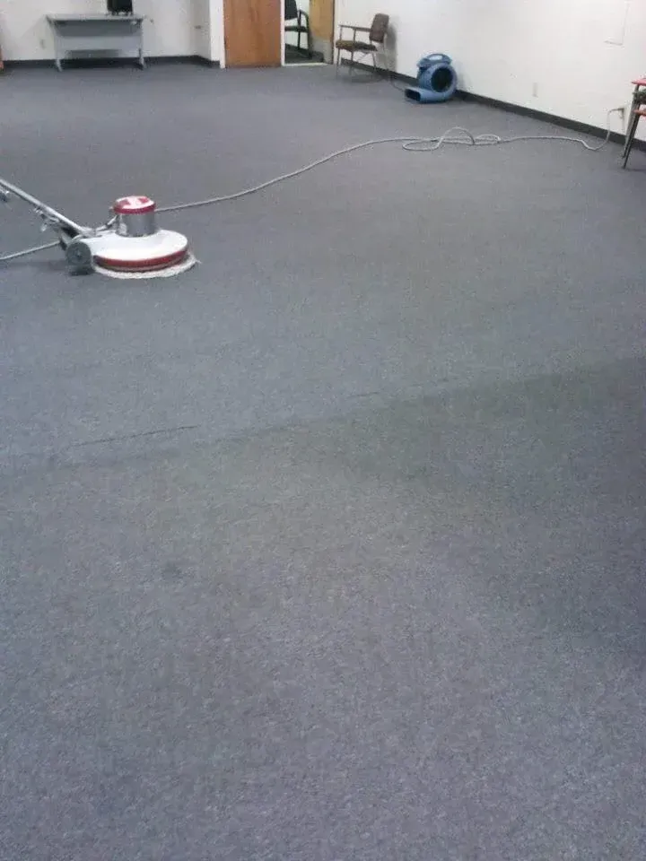 A floor buffer operates on grey office carpet, with a blue air mover in the background.