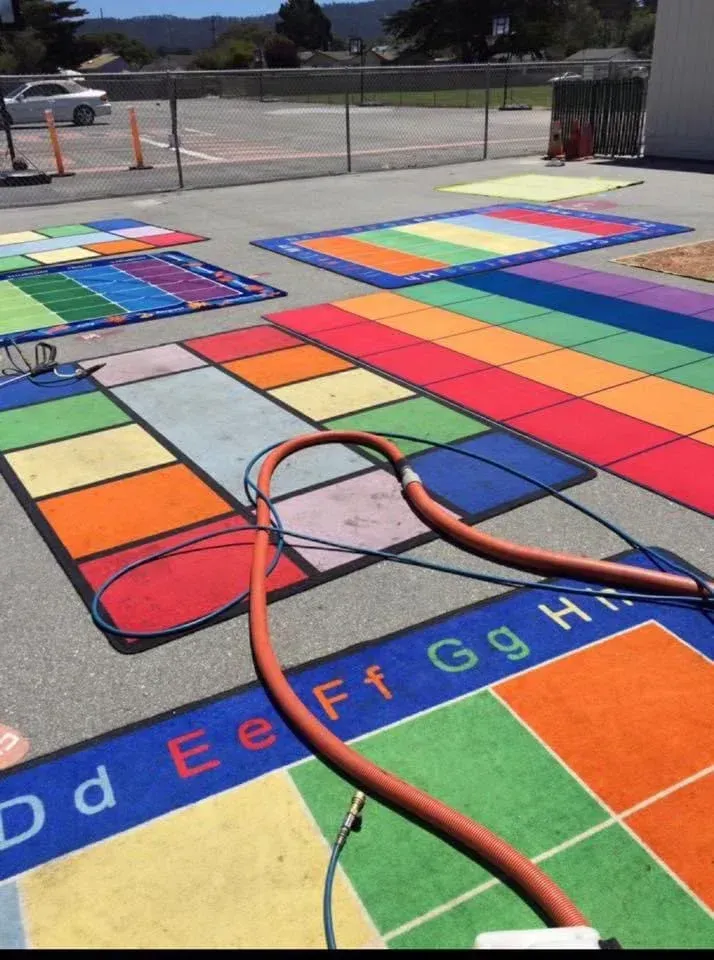 A colorful, grid-patterned educational mat lies on outdoor pavement, with a pressure washing hose snaking across it.