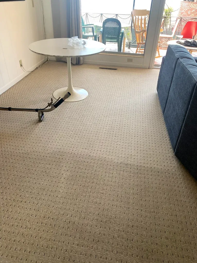 A room with beige patterned carpet, a white pedestal table, and a blue sofa, with a cleaning tool on the floor.