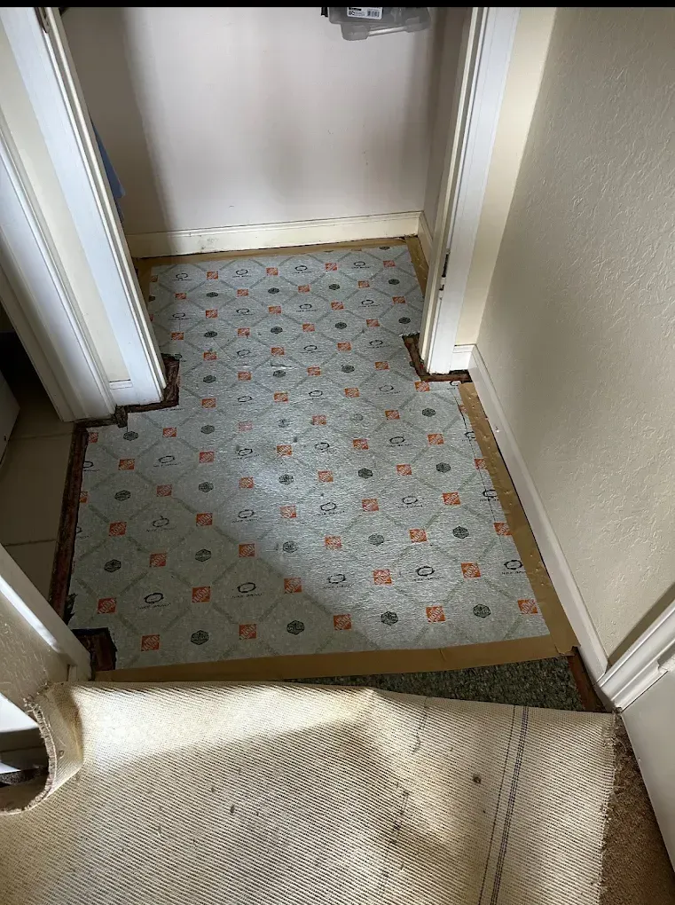 A light blue and white grid-patterned floor tile in an open closet, partially covered by beige carpet being pulled back.