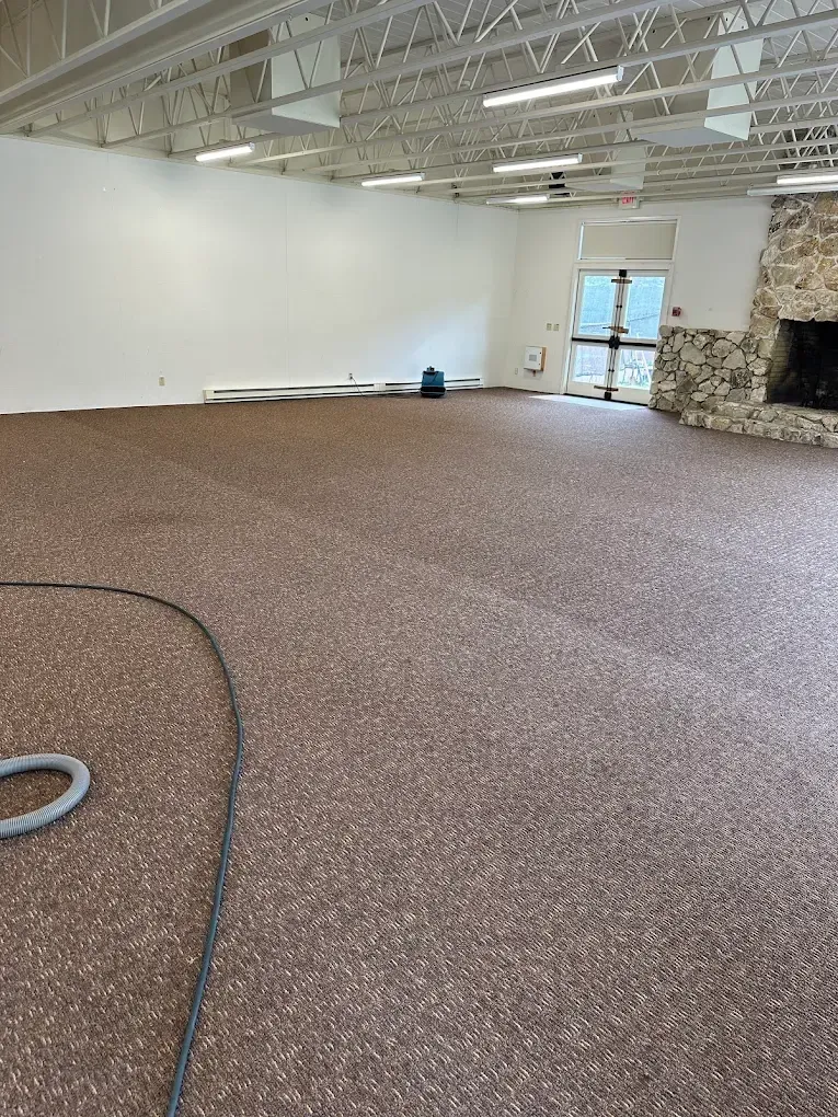 A spacious, empty room featuring speckled brown carpet, white walls, exposed ceiling trusses, and a stone fireplace.