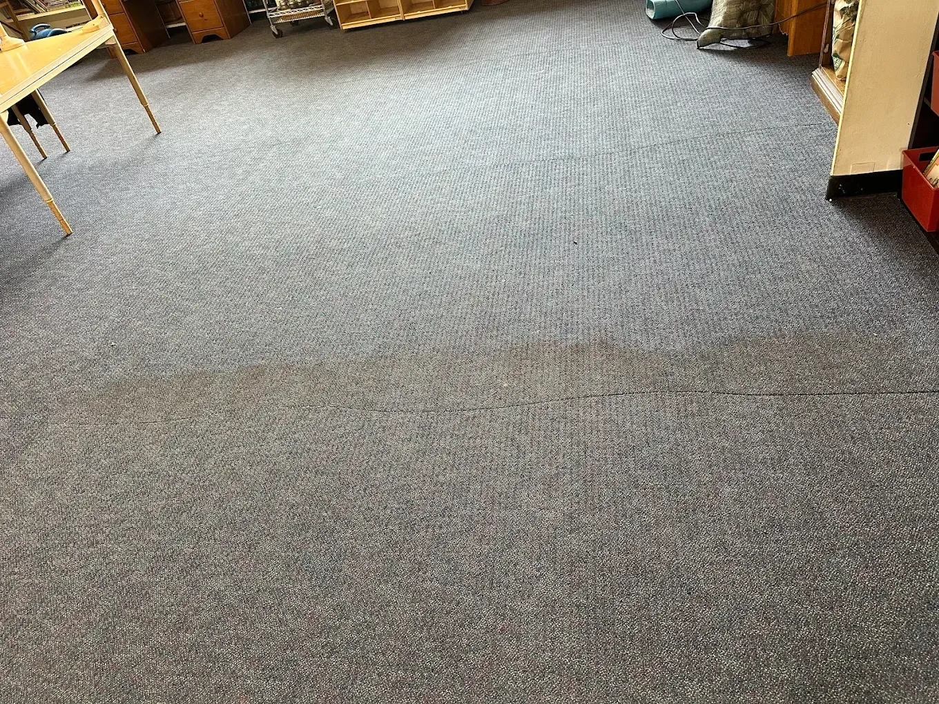 A wide view of a grey, textured carpeted floor in a room, with the legs of a wooden desk visible in the upper left corner.