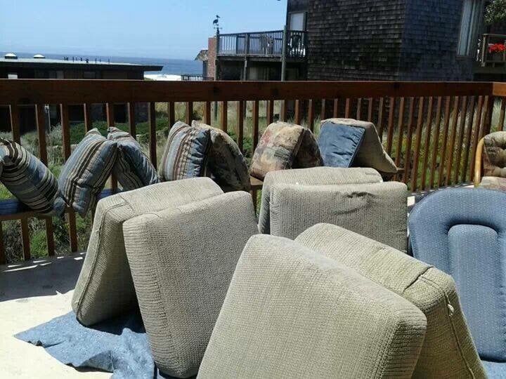 Several couch cushions and pillows propped up to dry on a wooden deck overlooking a coastal landscape.