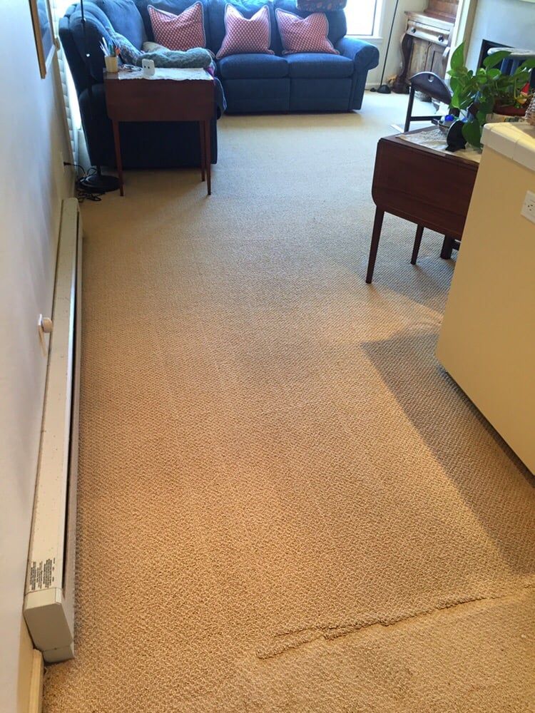 A living room with beige wall-to-wall carpet, a blue sofa, and a table. A metal baseboard heater runs along the left wall.