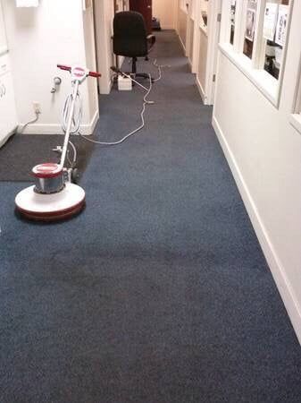 A floor buffer sits on a dark blue office carpet in a narrow hallway with white walls.