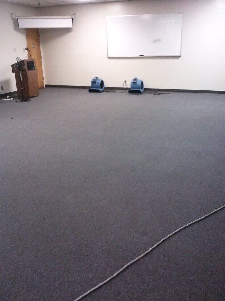 A room with gray carpet, a lectern, a whiteboard, and two blue floor-drying fans.
