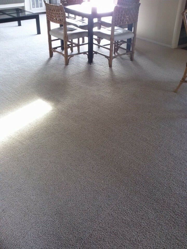 A dining table with four wicker chairs sits on a light-colored, speckled carpet in a brightly lit room.