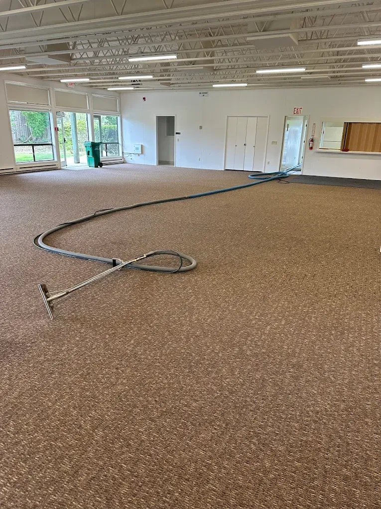 A carpet cleaning wand and long hose sit on a large, tan-colored carpet in an empty commercial space with white walls.