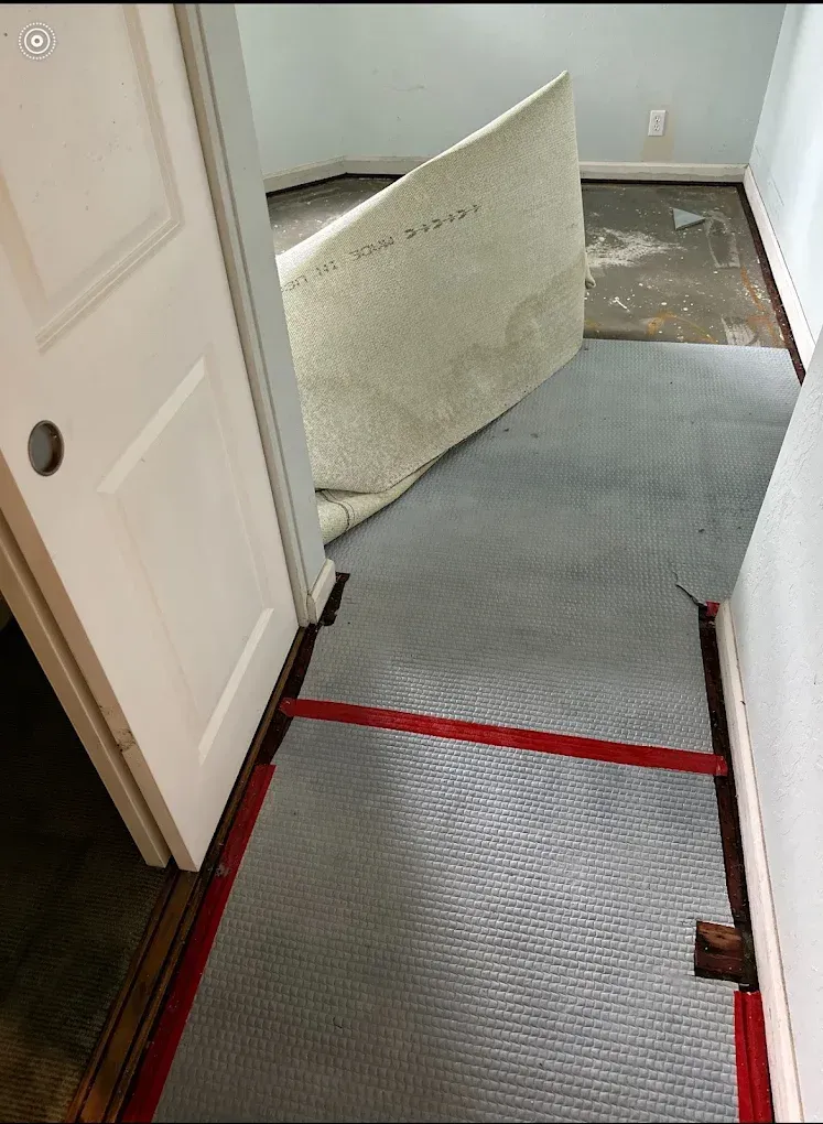 A room showing carpet being removed, revealing subflooring with red tape marking the floor surface.