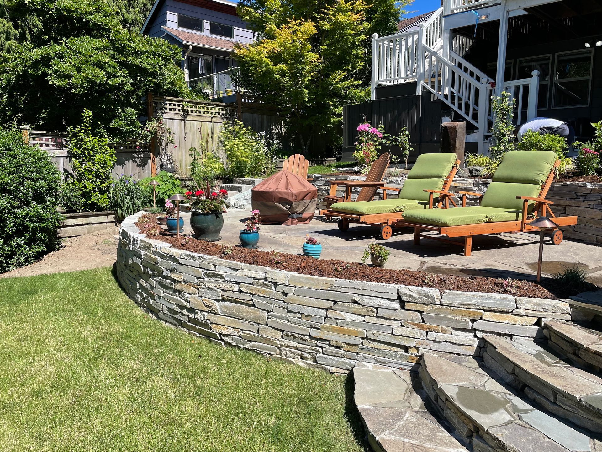 A stone wall surrounds a patio with lawn chairs and a fire pit.