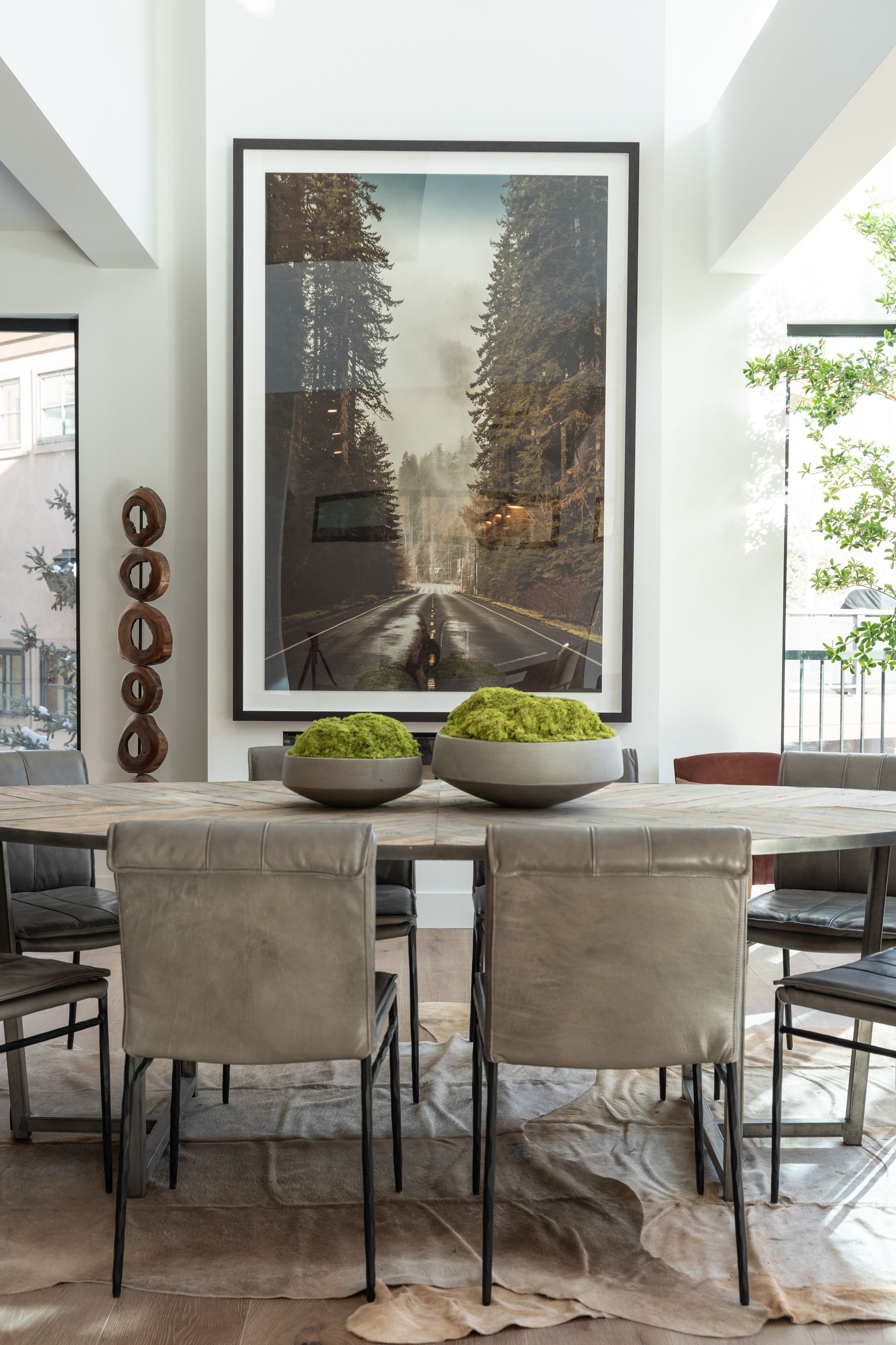 Dining room with large art piece of a street scene, table with two green plant bowls, and neutral-toned chairs and rug.