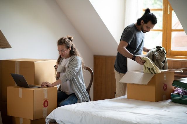 One person packing moving boxes on a bed while another person sits in a chair using a moving box as a desk for their laptop