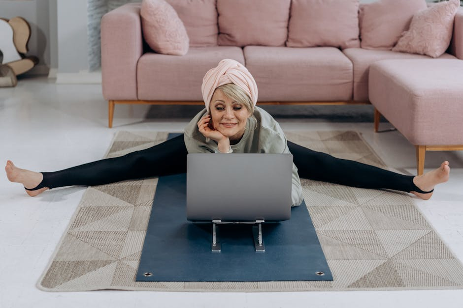 An elderly woman is doing splits on a yoga mat while using a laptop.