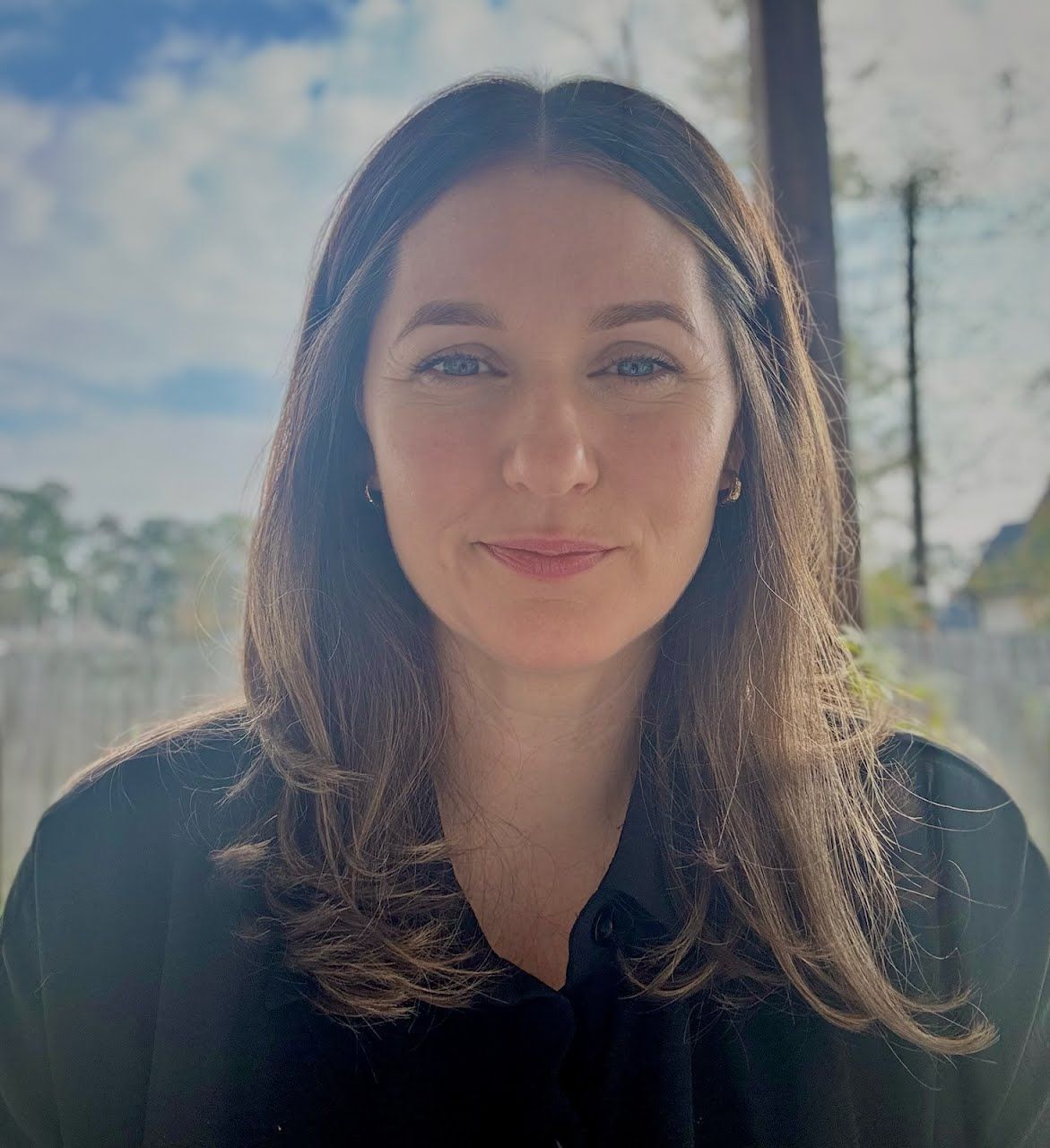 Woman with long brown hair looks at the camera. She's wearing a dark shirt. Outdoors with sky and trees.