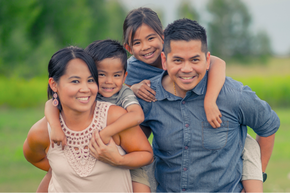 A picture of a family with two kids on the backs on the parents with green grass behind them.