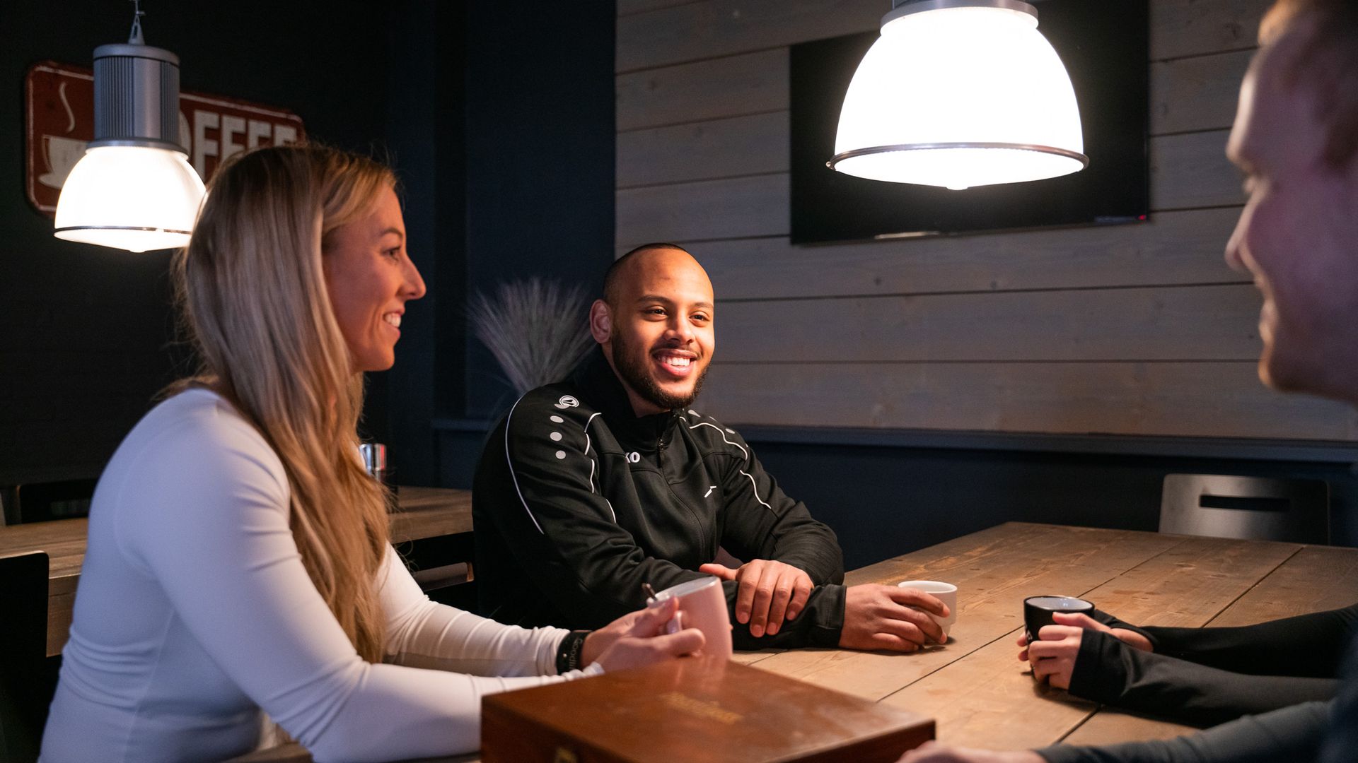 Drie mensen zitten aan een houten tafel in een café, lachend en met elkaar in gesprek.