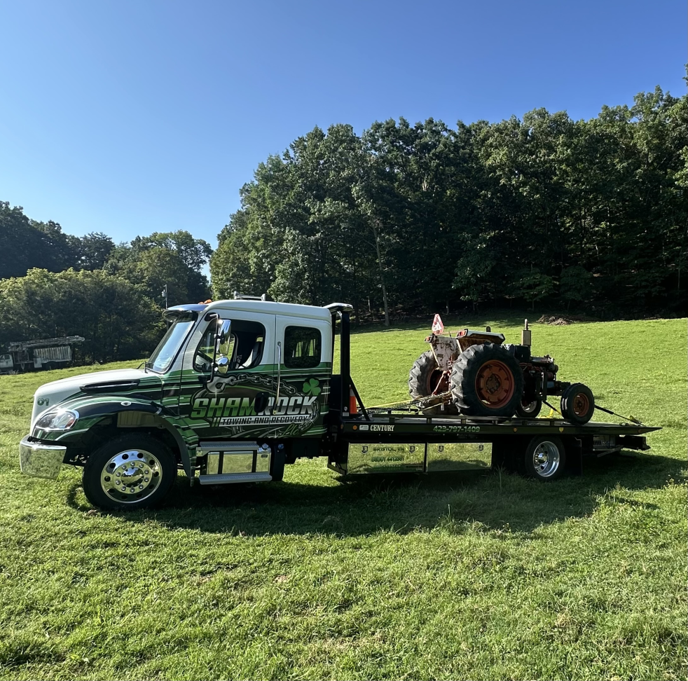Tow truck carrying a small tractor on a flatbed in a sunny grassy field