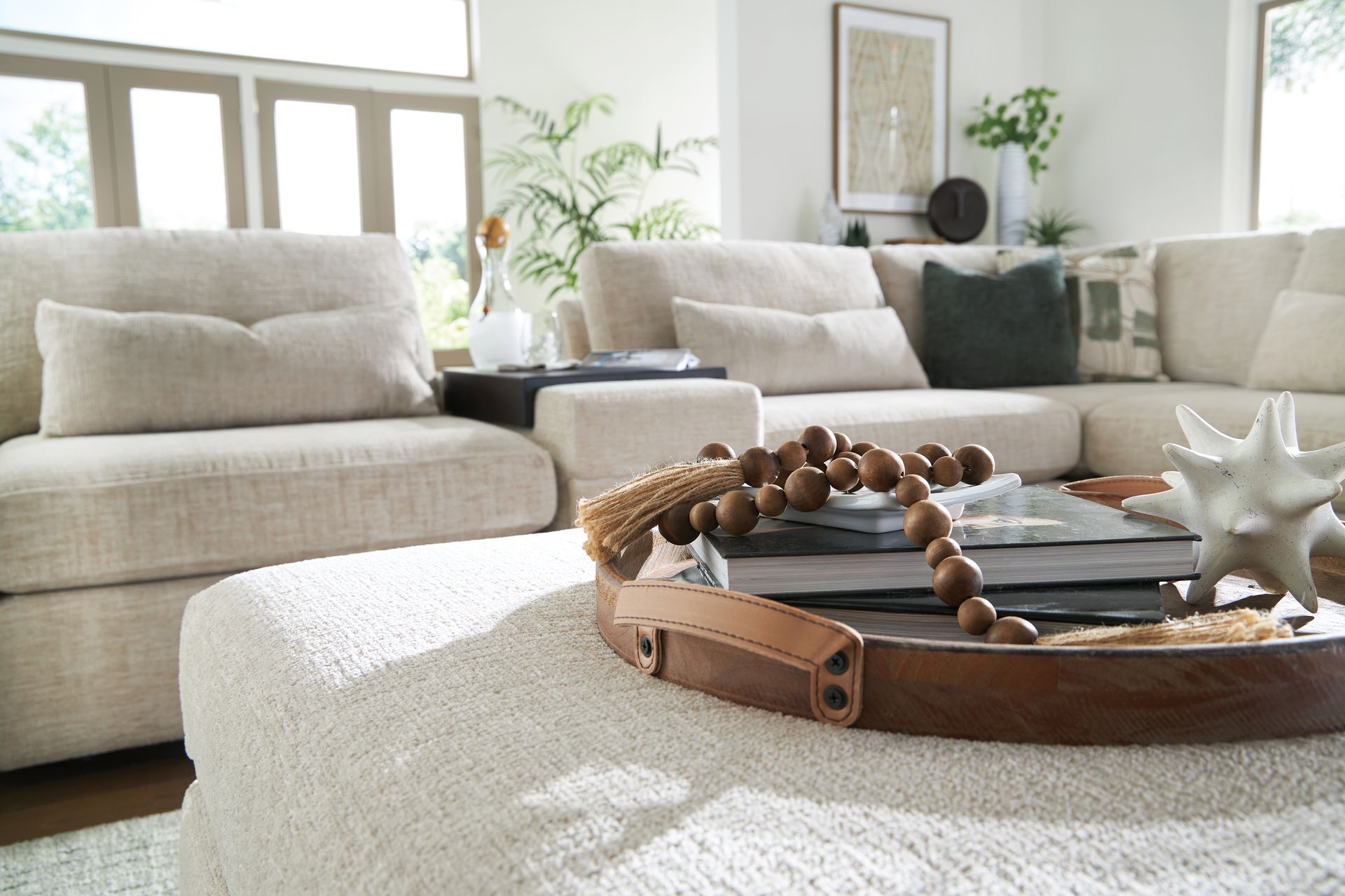 Living room with beige sectional sofa and ottoman, a wooden tray with beads, and decor.