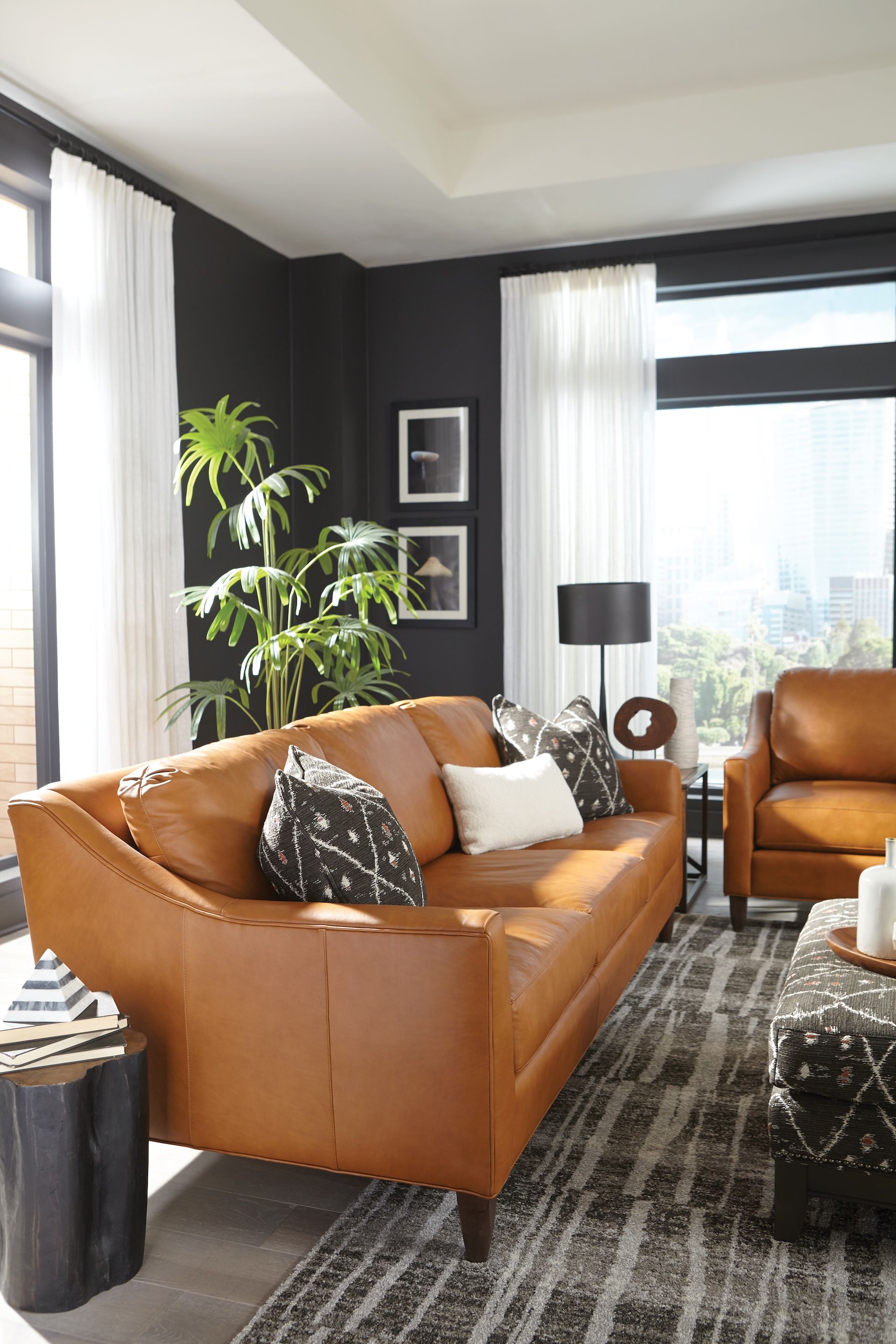 Brown leather sofa and chair in a modern living room with dark walls and large windows.
