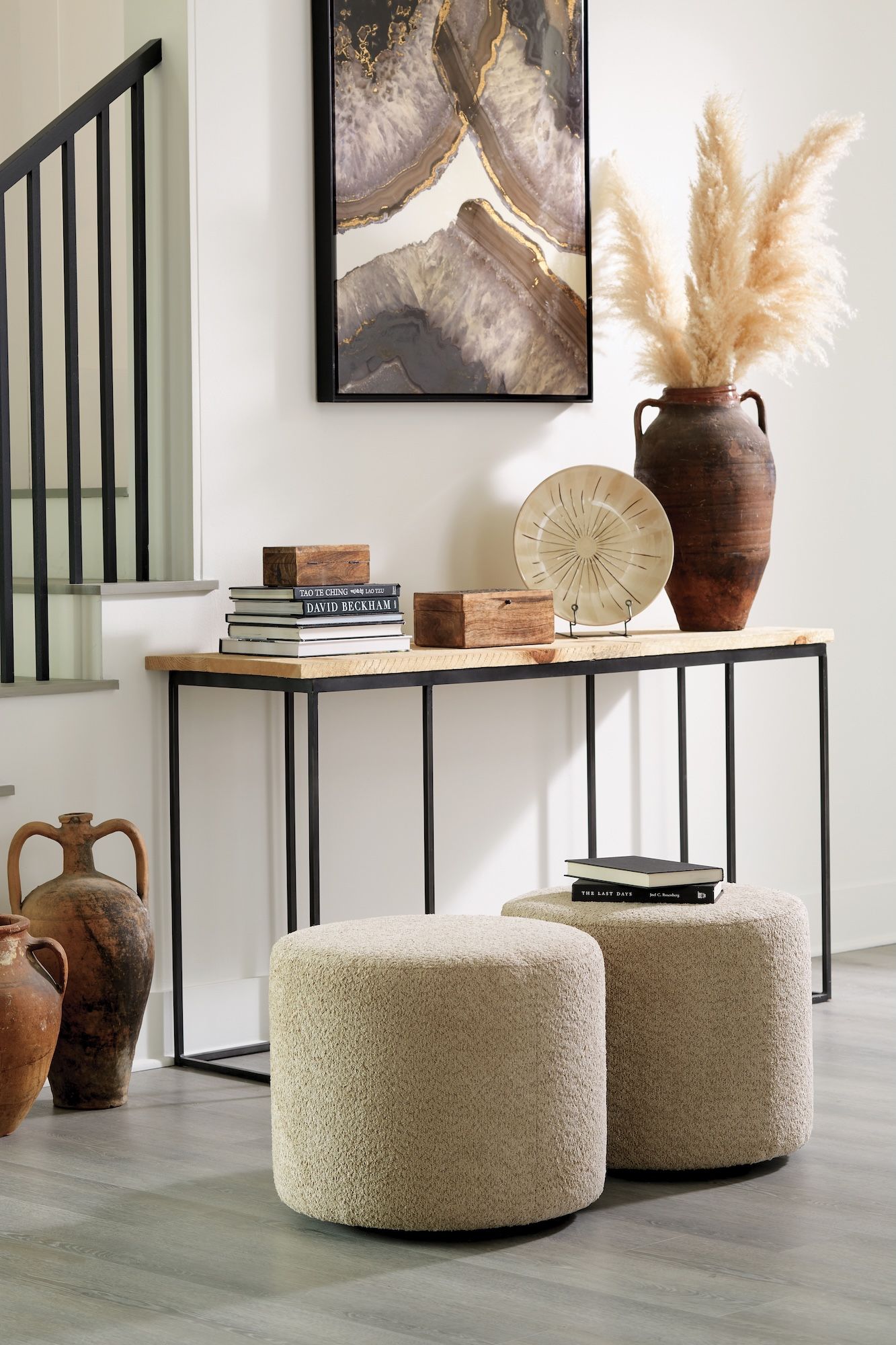 A console table with decor, including a vase with pampas grass, two ottomans, and wall art.