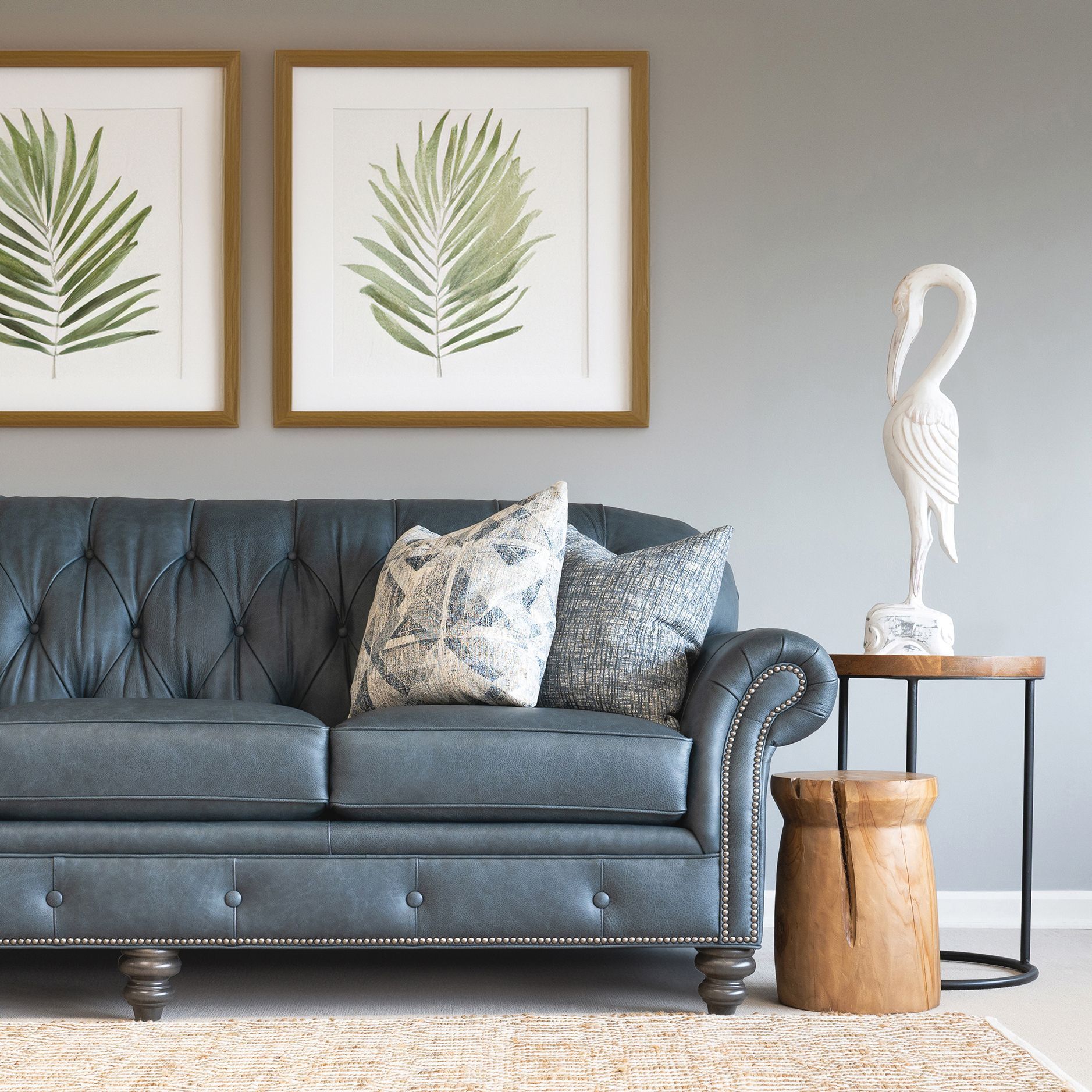 Blue tufted sofa with pillows, art, wooden side table with bird statue and stool, beige rug, gray wall.