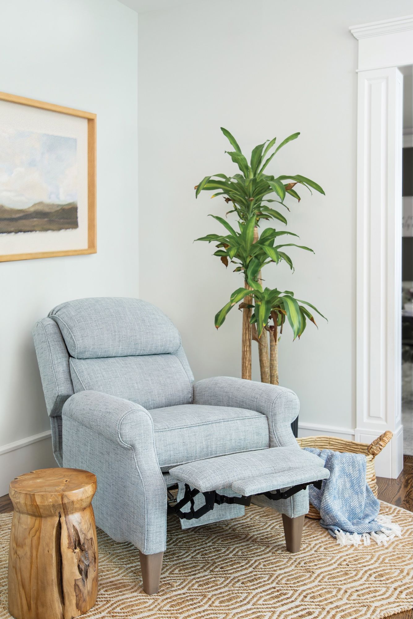 Light blue recliner with footrest extended, small wooden side table, large plant, painting.