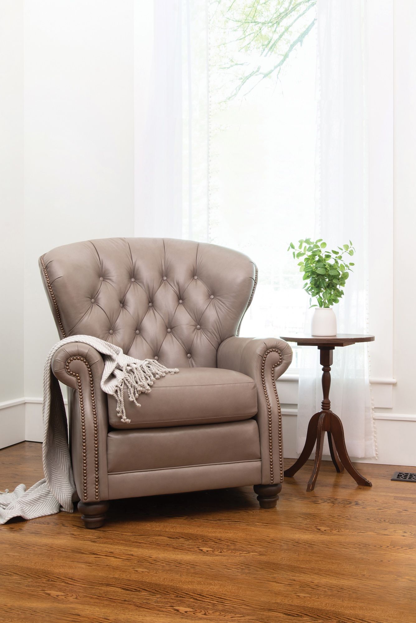 Tufted grey leather armchair with a blanket next to a small wooden side table with greenery, by a bright window.