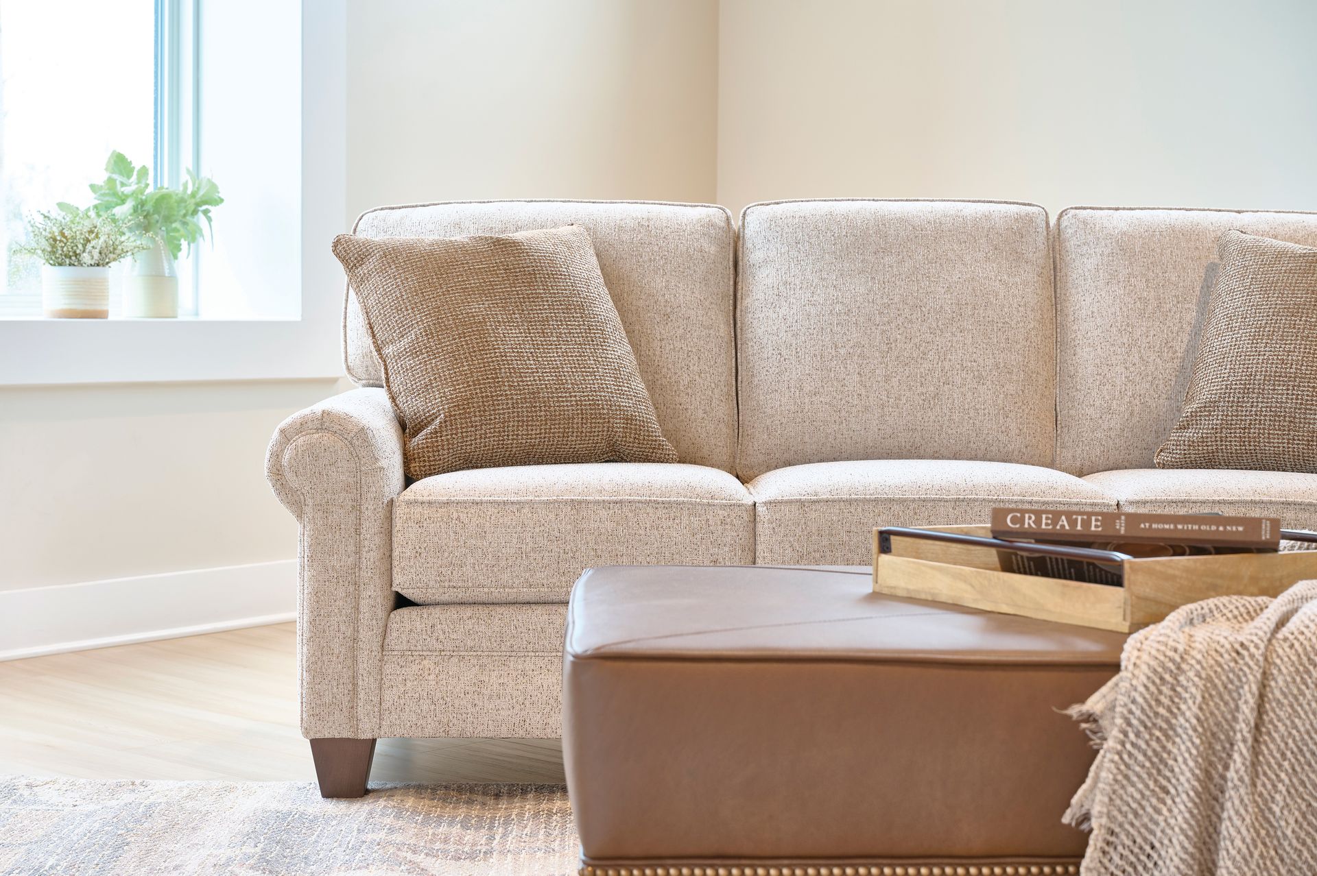 Beige sofa with brown accent pillows, leather ottoman, and tray of books in a sunlit room.