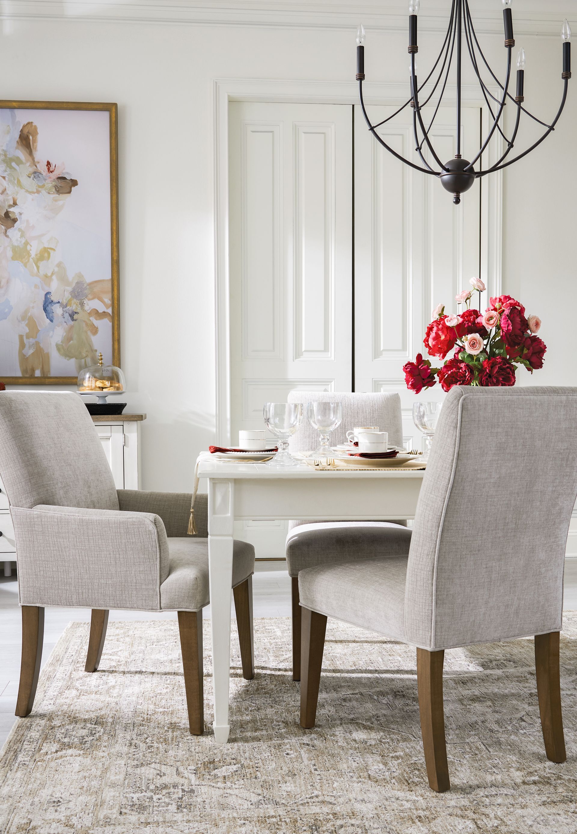 Elegant dining room with white table, chairs, and chandelier. Artwork and flowers add color to the light space.