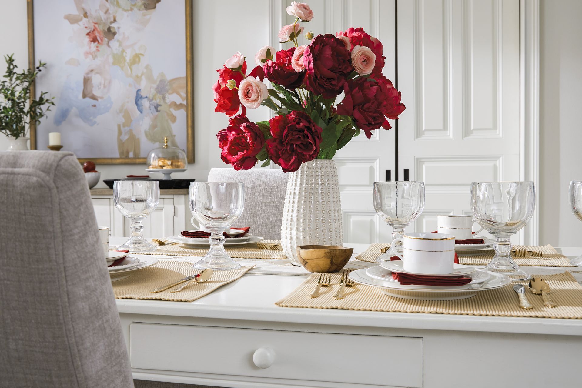 Elegant dining table set with red flowers in a vase, place settings, and artwork.