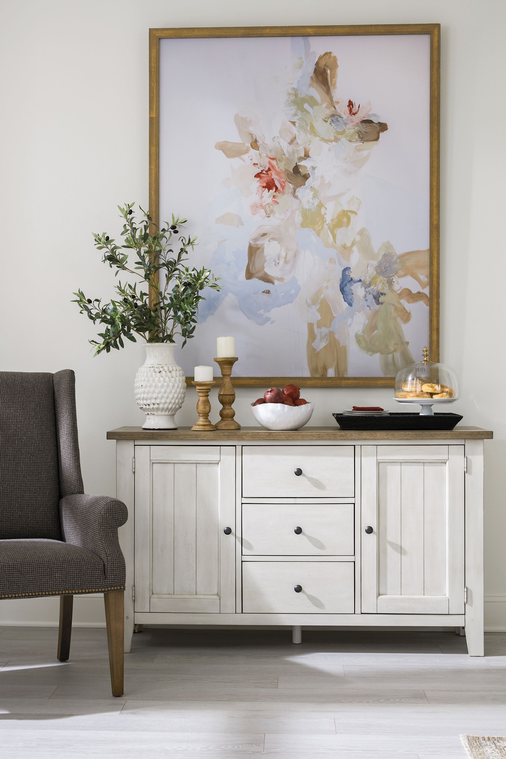 White buffet with art, vase of branches, chair, and candles against a white wall.