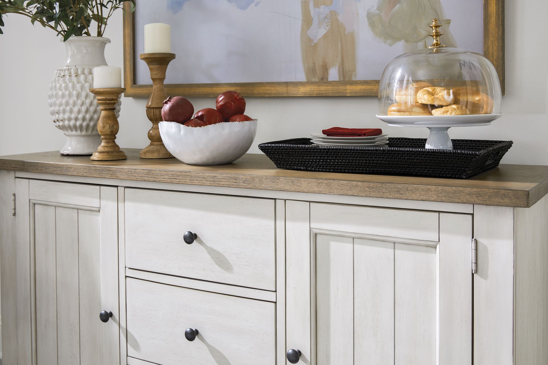 White buffet with decor: candles, fruit bowl, cookies, and framed artwork.