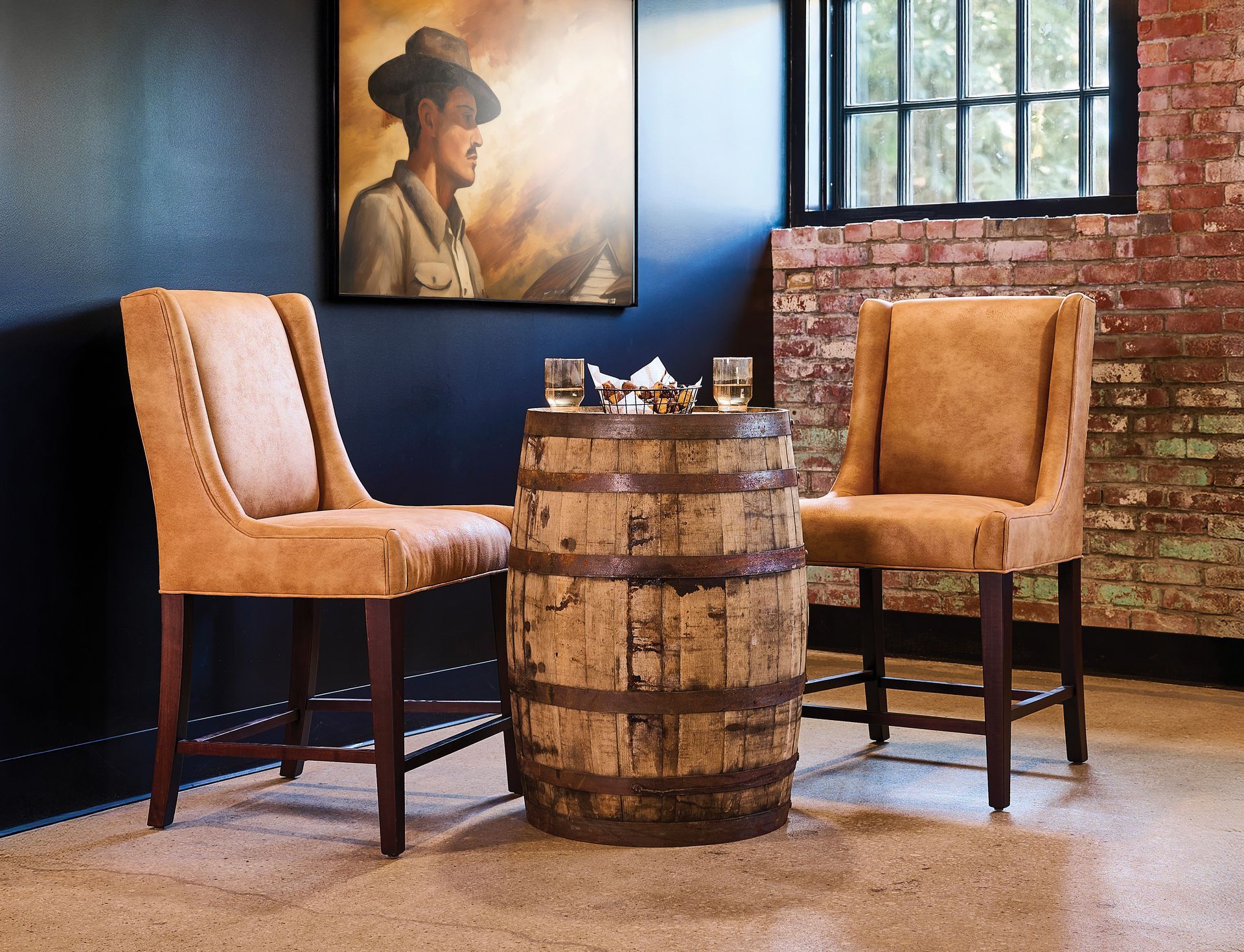 Two tan chairs flank a barrel table in a room with a painting and brick wall.