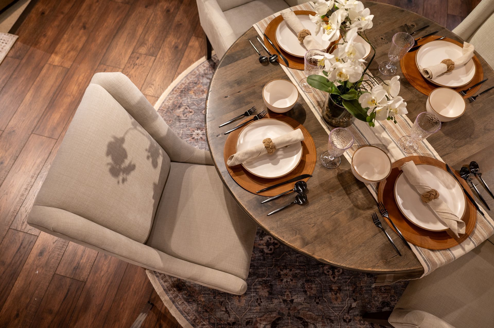 Round wooden dining table set for four with white flowers, beige plates, and silverware.