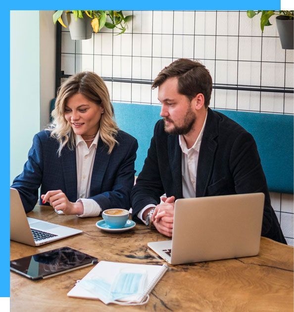 Two professionals, a man and woman, looking at laptop screens at a cafe table.