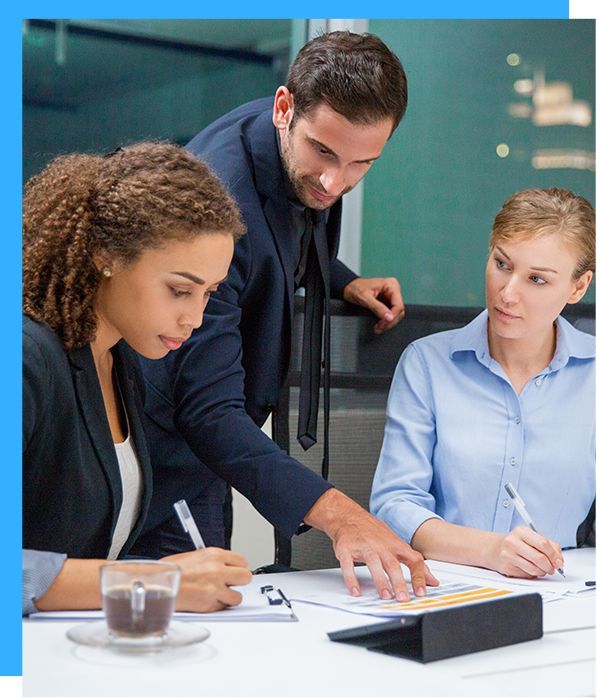 Business team in meeting, reviewing documents. Man points, women write, coffee cup visible.
