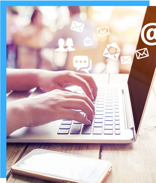 Person typing on laptop, social media icons floating above, phone below on wooden table.