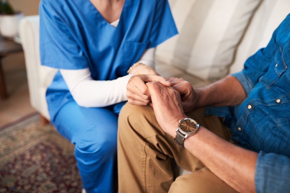 Nurse in blue scrubs holds the hands of an elderly man seated on a couch.