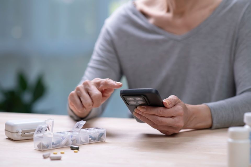 Person using phone next to pill organizer and medication bottles, indoors.