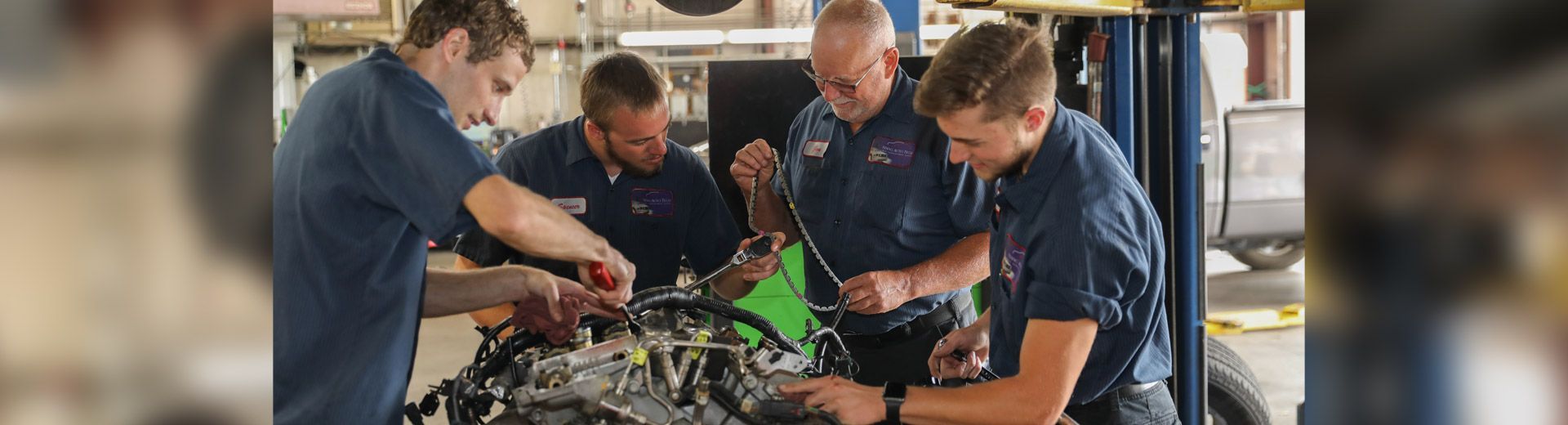 Four men in blue shirts work together on an engine in a garage setting. They appear to be mechanics. | Stang Auto Tech