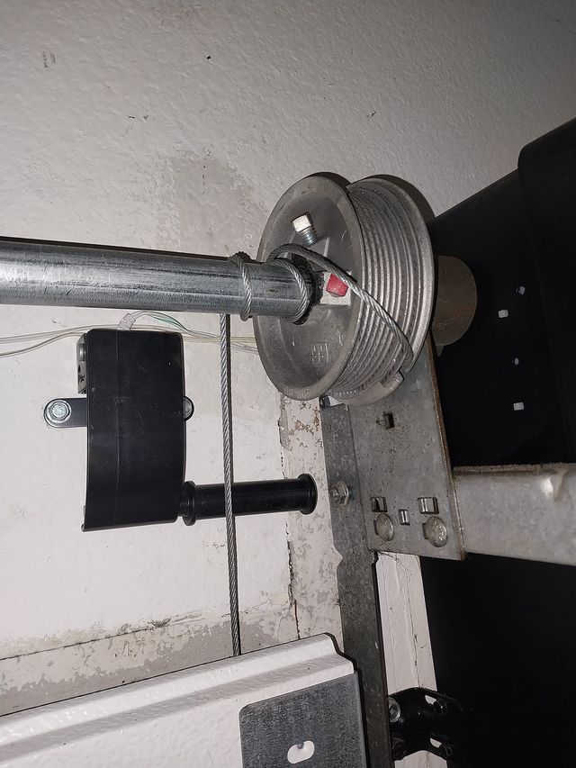 Metal cable drum and pulley mounted on a wall beside a black strap and chain mechanism