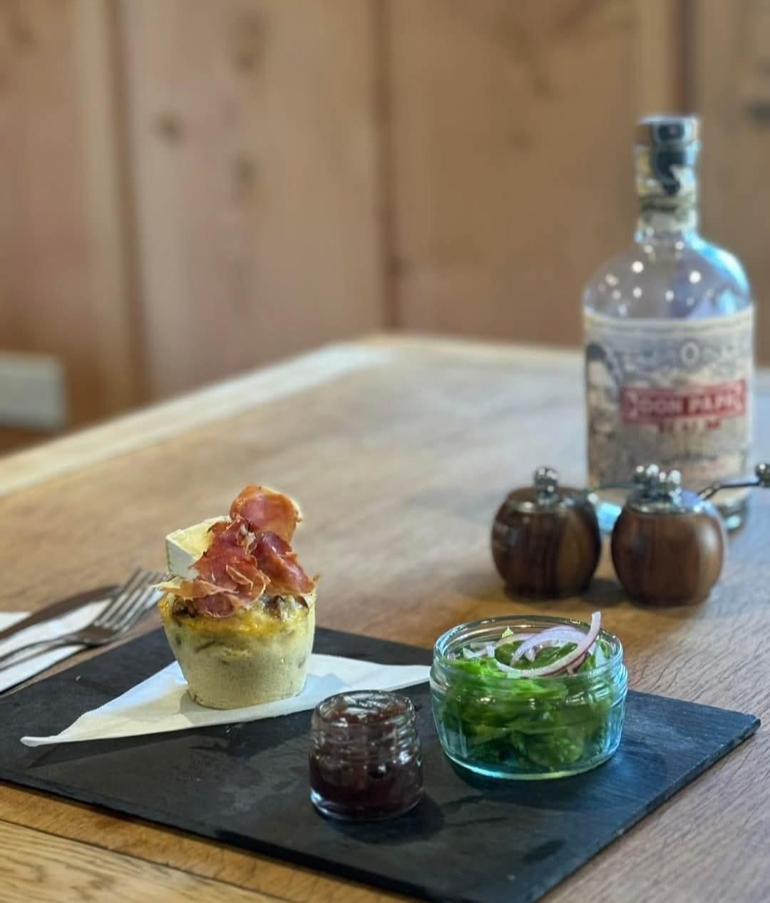 Appetizer on a slate board: quiche, jam, green salad with onion, salt and pepper shakers, and a bottle on a wooden table.
