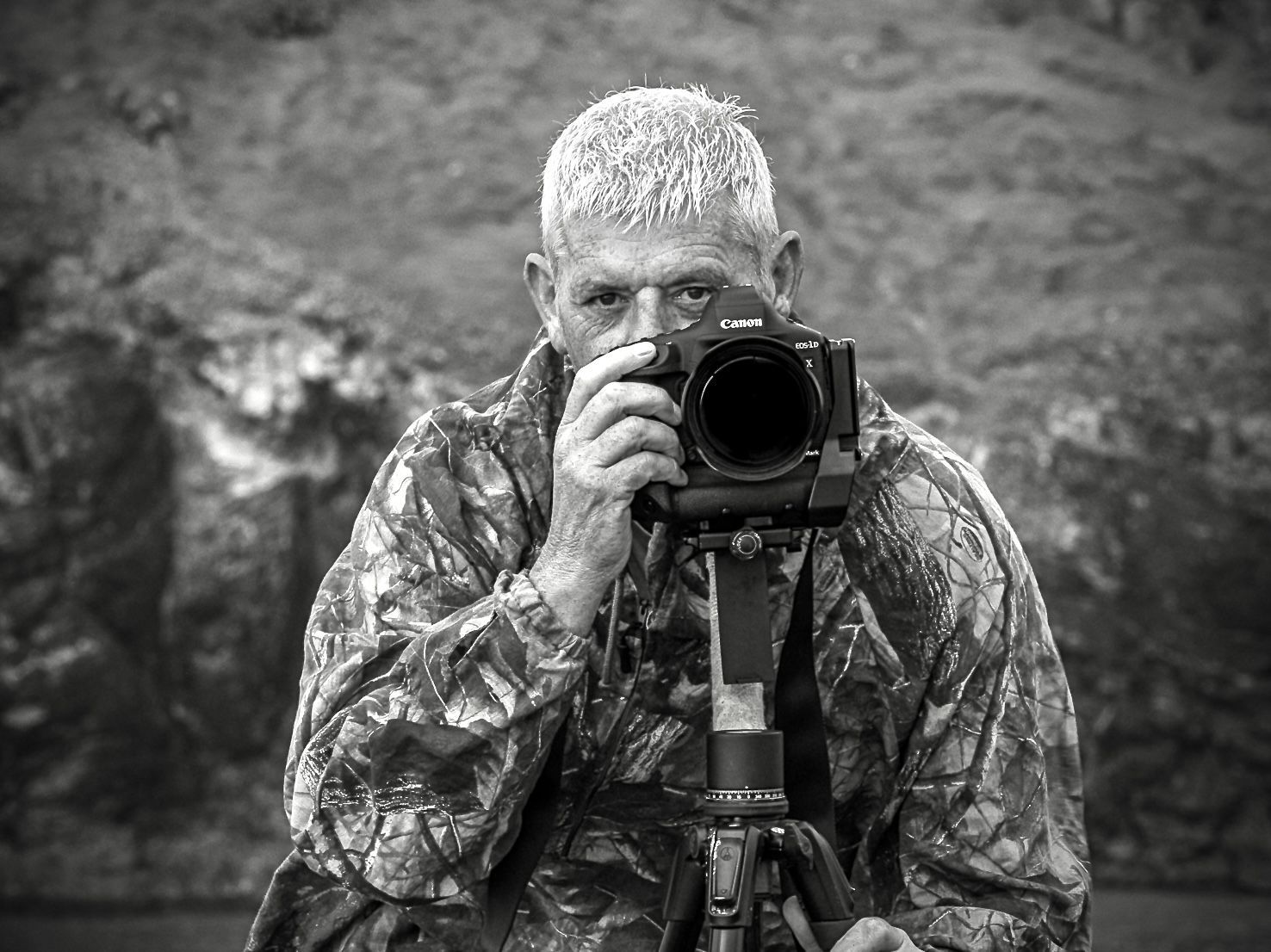 Photographer in camouflage, holding a camera on a tripod, looking through the lens.