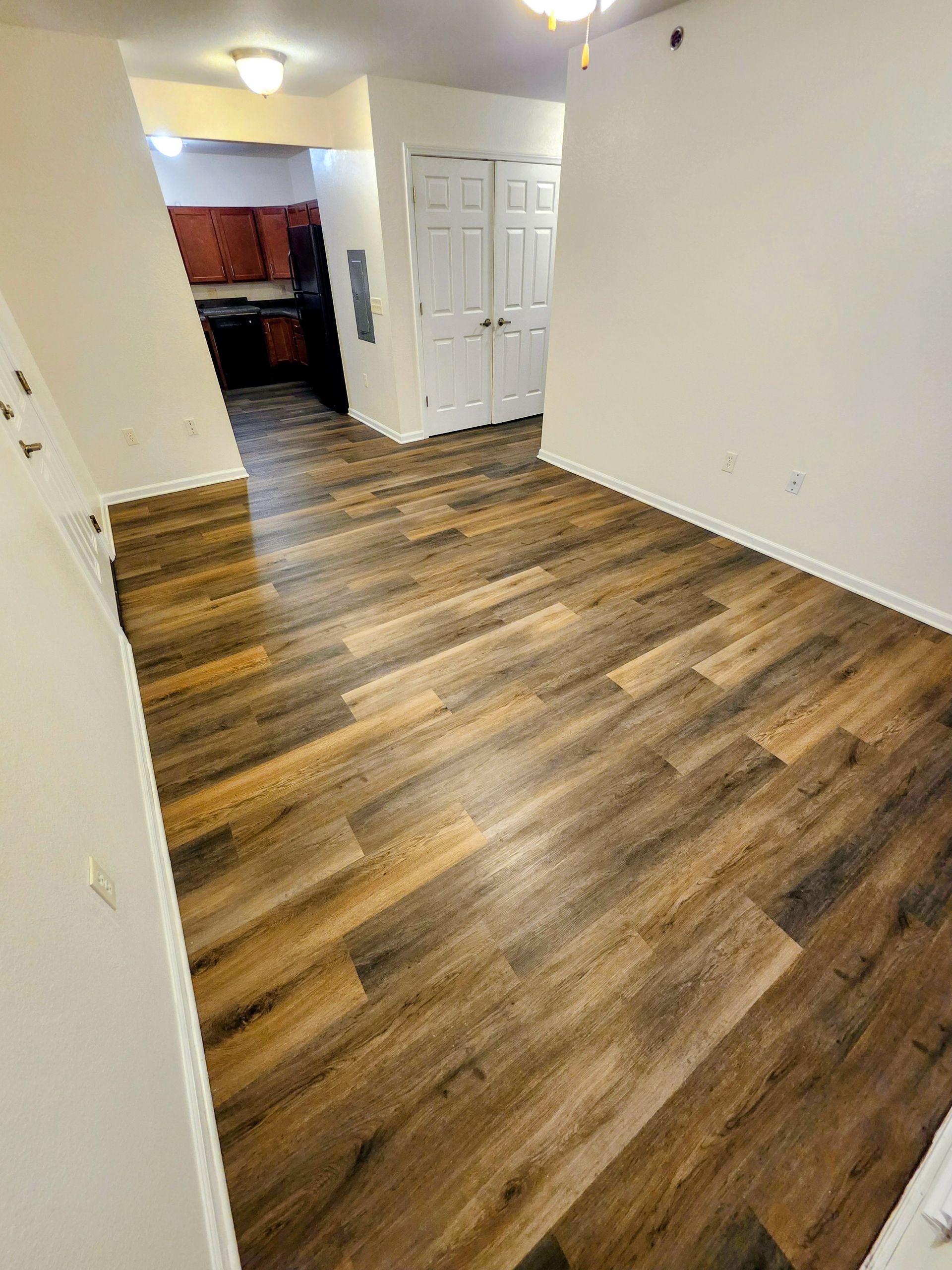 Empty apartment room with wood-look flooring, white walls, and a hallway leading to a kitchen.