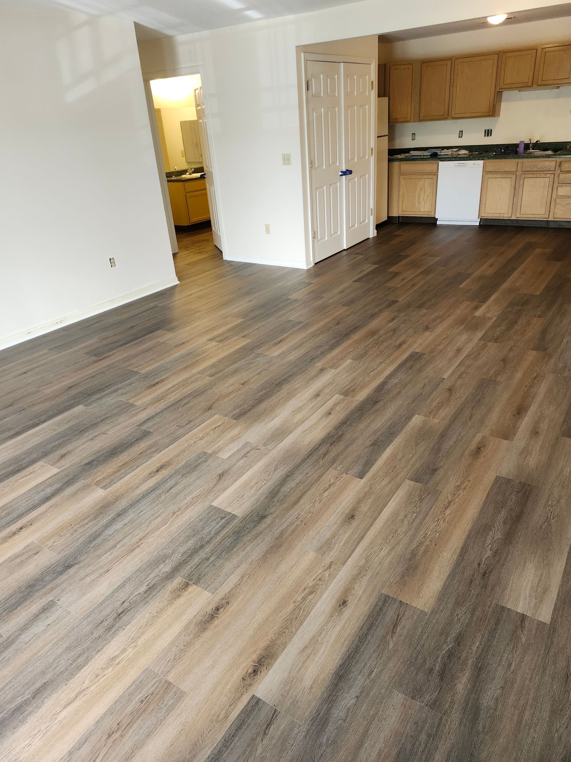 Empty apartment living area with wood-look flooring, white walls, and a small kitchen in the background