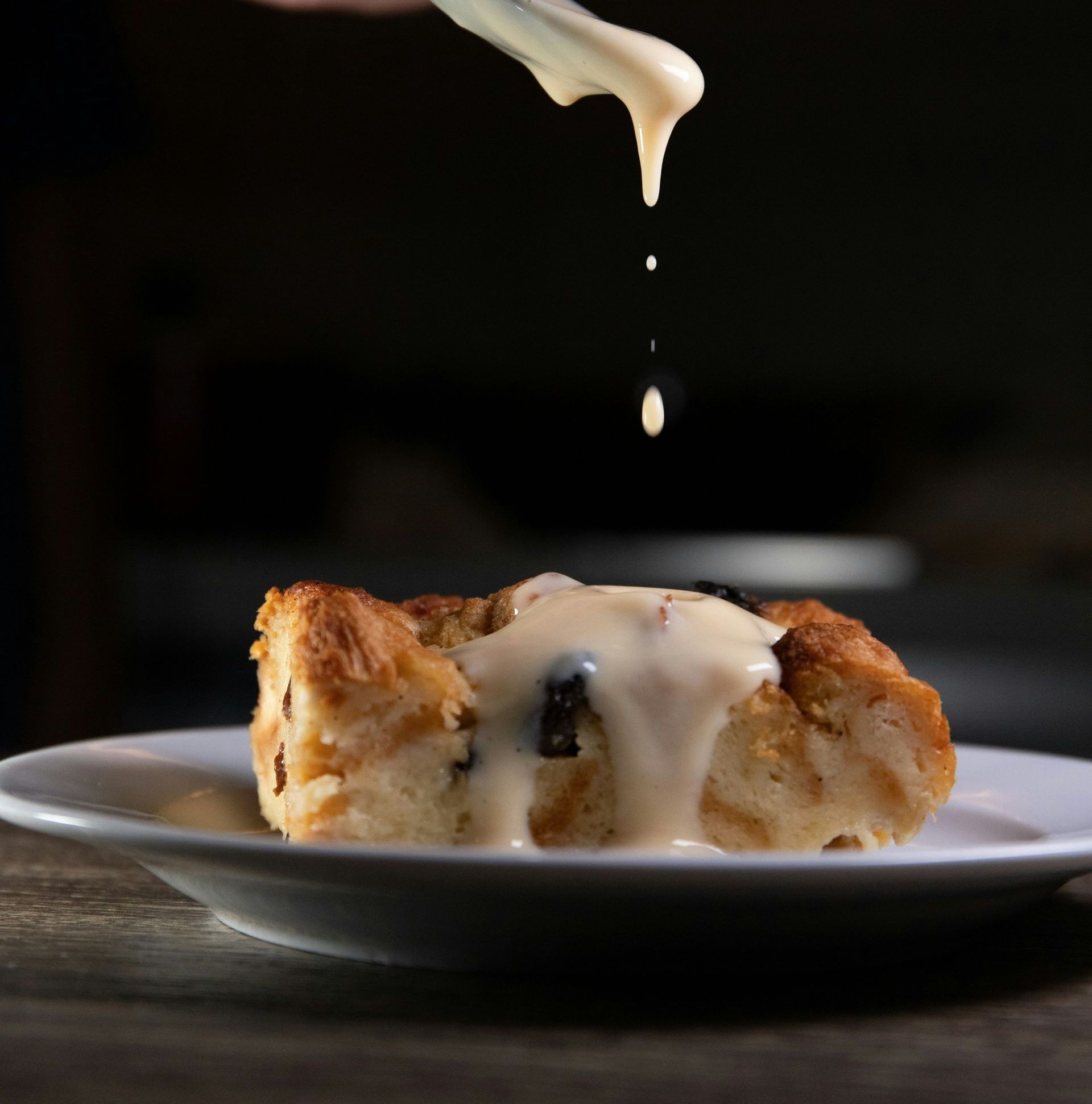 A piece of bread pudding on a white plate with icing being poured on it
