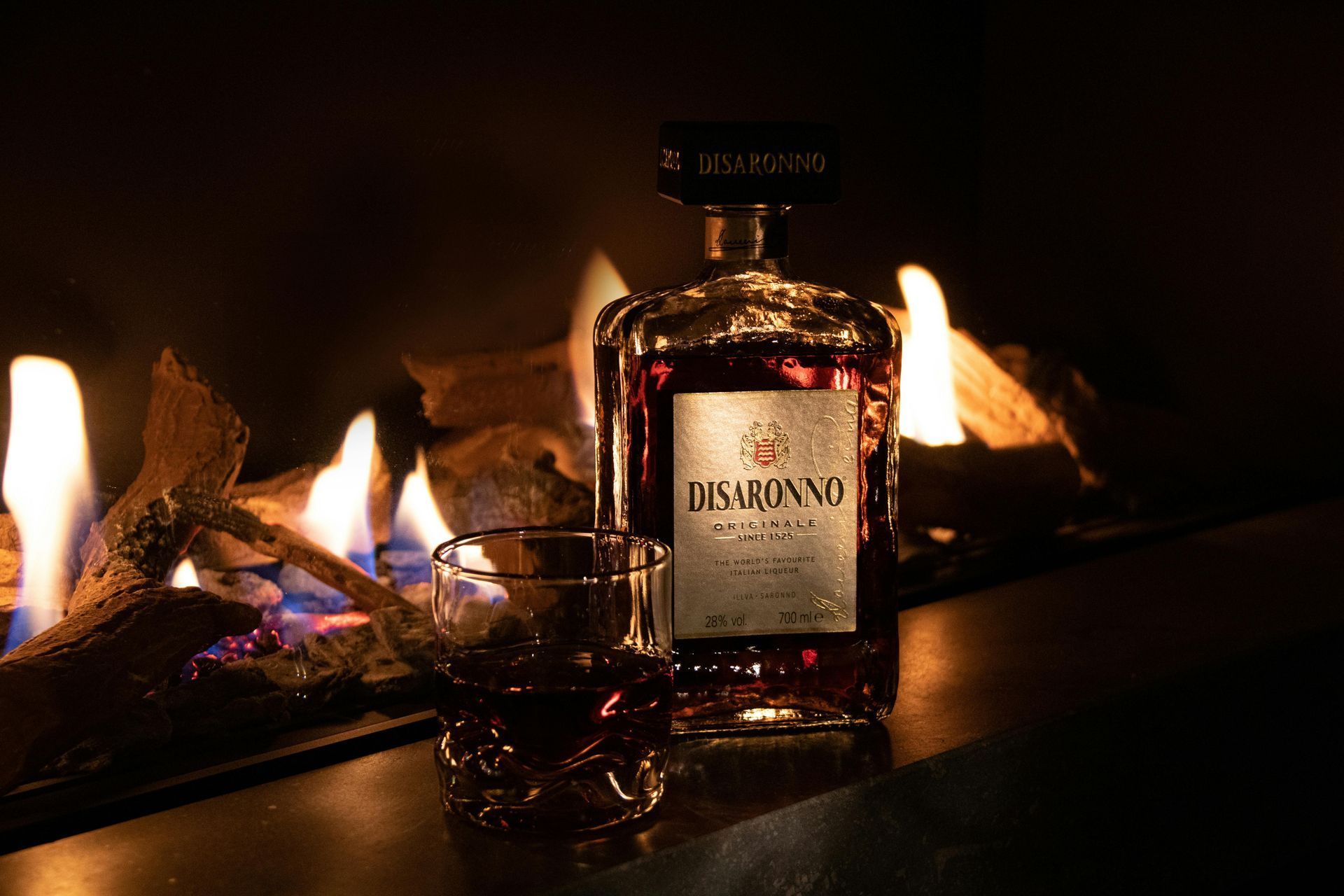 A bottle of disaronno next to a glass of whiskey in front of a fireplace.