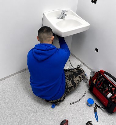 Technician in blue hoodie repairing plumbing under a wall-mounted sink in a small bathroom.